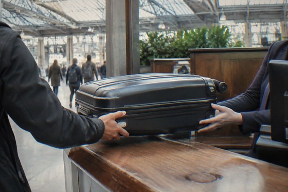 A traveller handing a suitcase across a staffed service counter in a bright train hall