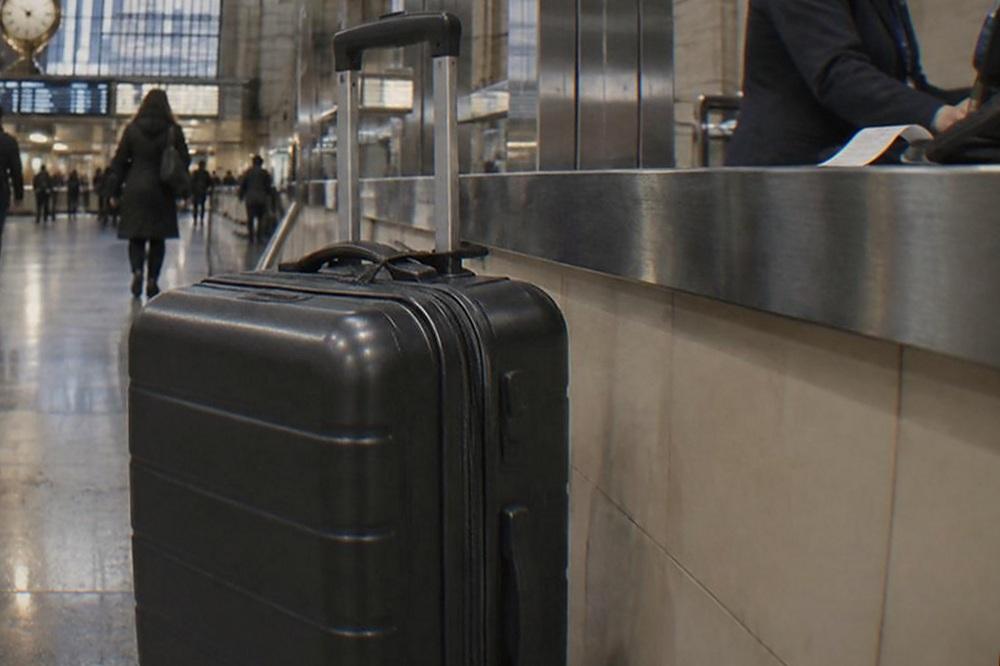 A staffed baggage counter in a train station with a rolling suitcase waiting beside it