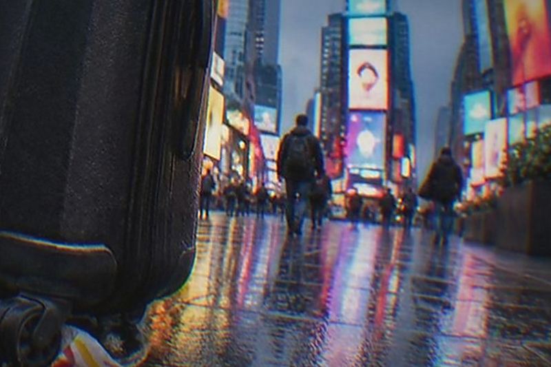 A traveller sets a suitcase down at a Manhattan street corner within sight of a famous New York landmark, about to drop the bag at a nearby partner counter.