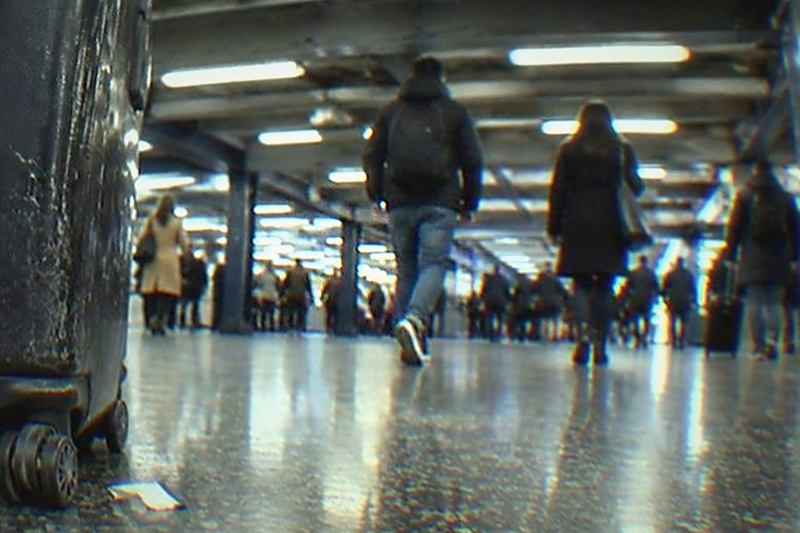 A suitcase waits beside a traveller on the concourse of a busy New York transport hub, where passengers pass in the background and a staffed bag drop sits within sight.