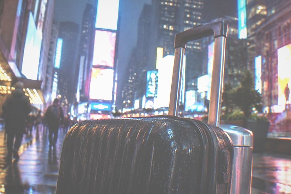 Late night street corner in a bright theatre district with lit marquees and a closed suitcase waiting at the kerb