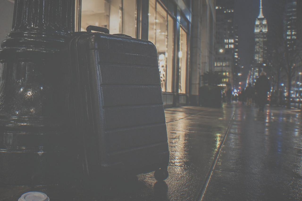Empty Manhattan street at night with glowing storefront lights and a closed suitcase resting against a lamppost