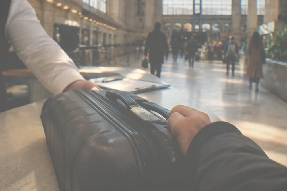 A traveller handing a suitcase across a service counter while a staff member checks a clipboard