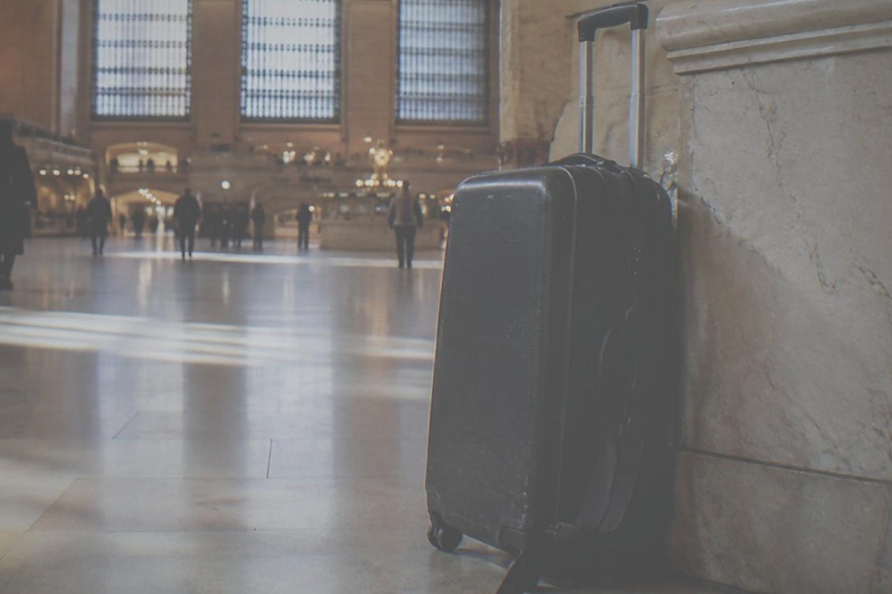Empty train hall concourse with sunlight through tall windows and a single suitcase standing by a pillar