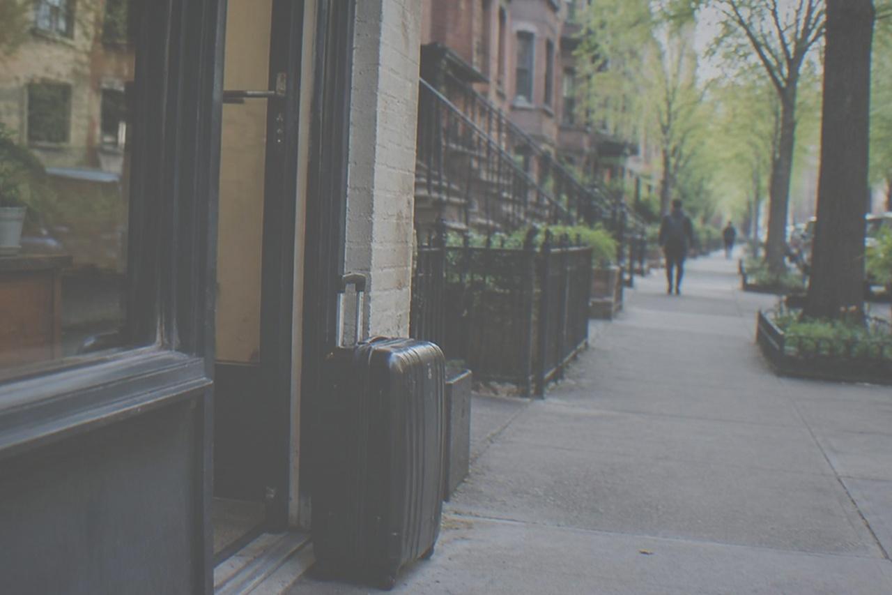 A quiet Manhattan sidewalk outside a small shop with a suitcase parked beside the door