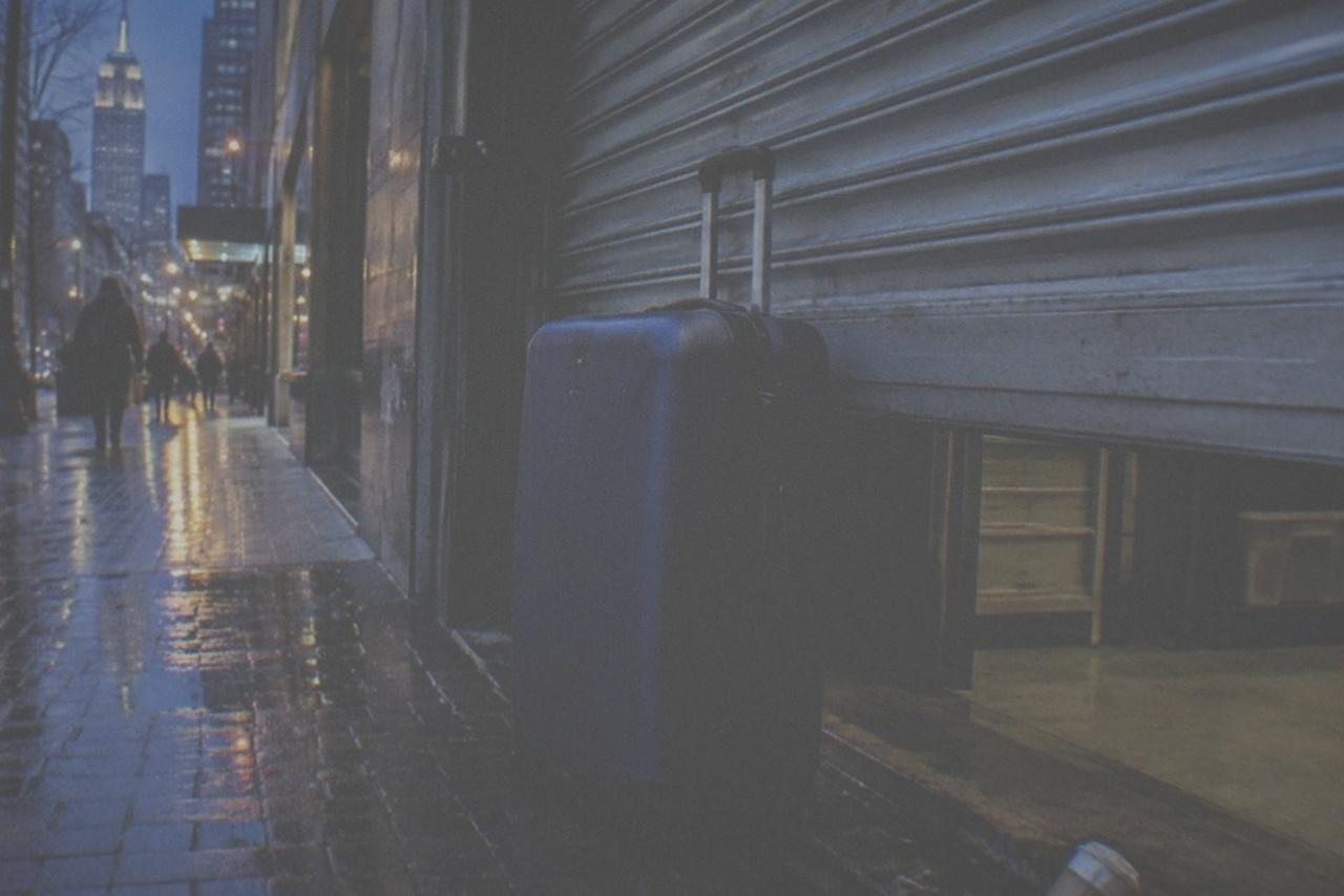A closed storage shop front on a quiet Manhattan side street at dusk, shutter halfway down, a single suitcase parked beside the door