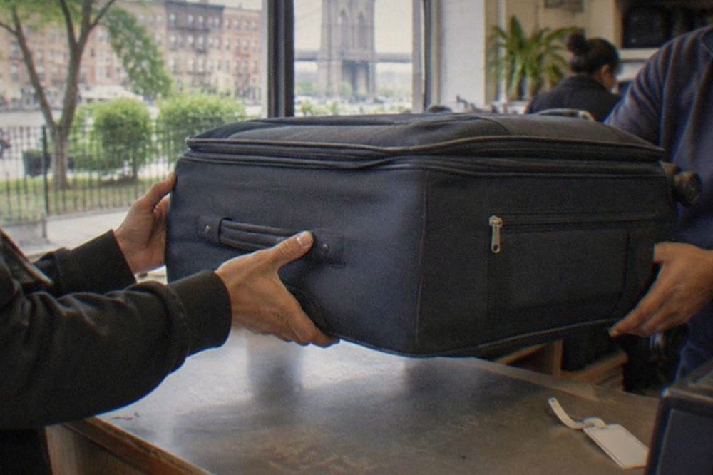 Traveller handing a large suitcase to a shopkeeper across a counter in a bright store