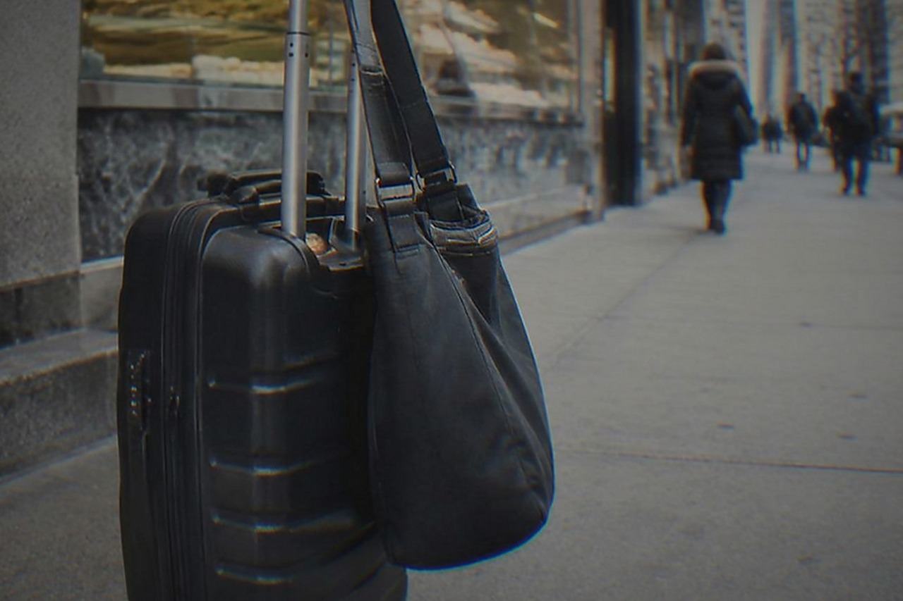 A traveller pauses on a New York City sidewalk with a wheeled suitcase and a shoulder bag, about to carry them into a shop and hand them to the attendant at a staffed storage counter.